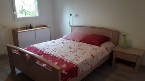 A bedroom with a wooden bed, a nightstand, a bedside table, a window with a view of greenery, and a wall-mounted lamp. - Appartement salon cuisine chambre jardin. Sécurisé. Neuf. hygiène +++ (la Roche sur Foron)