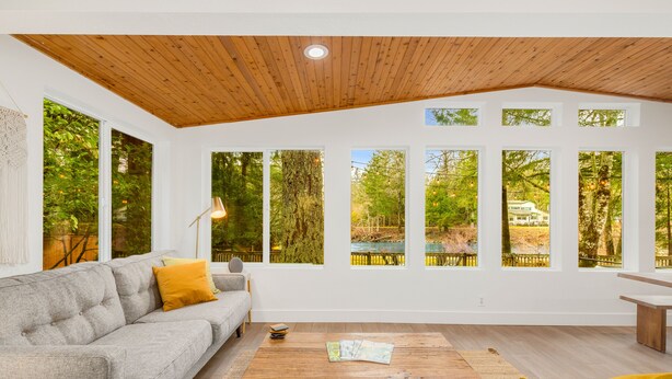 A modern living room with a grey sofa, wooden coffee table, and large windows offering a view of a forested area.