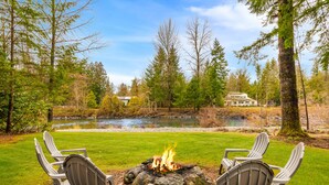 A fire pit with chairs in a grassy area with a river and trees in the background.