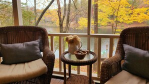 A wicker chair with a cushion, a small table with a vase of flowers, and a view of autumn trees through a screened window.