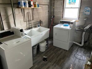 Une buanderie équipée d’un lave-linge, d’un évier et d’un sèche-linge. On y trouve des produits de nettoyage sur une étagère, ainsi qu’une fenêtre donnant sur l’extérieur.