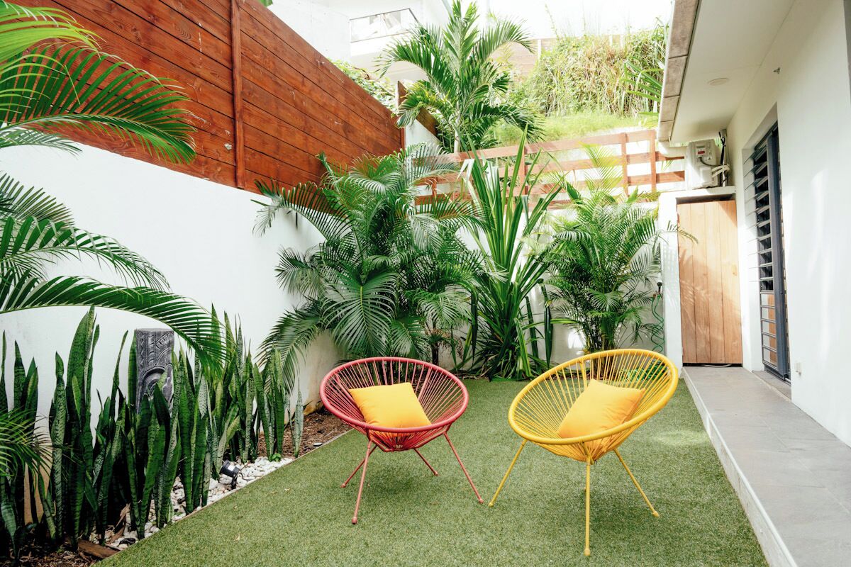 A small patio area with artificial grass, two wicker chairs with cushions, and various tropical plants.