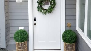 Porta de entrada com grinalda, duas plantas em vasos, tapete e luminária na parede.