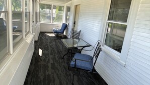Une terrasse couverte avec une table et des chaises en verre, des murs recouverts de panneaux blancs et de grandes fenêtres.