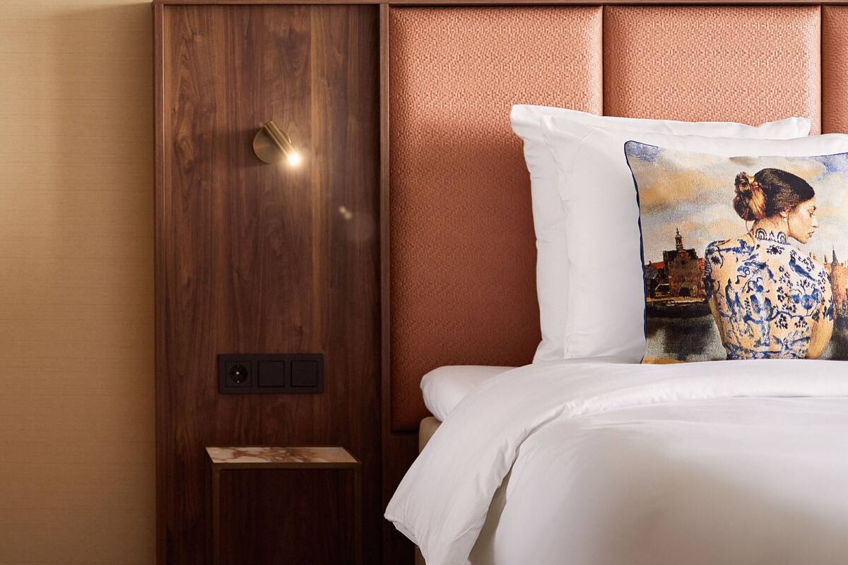 Een hotelkamer met een houten garderobe, een kleine tafel, een bed met witte beddengoed en een kussen met een schilderij.
