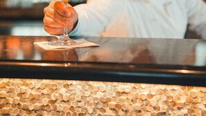A person holding a cocktail glass at a bar with a wooden hexagonal pattern floor and shelves with bottles in the background.