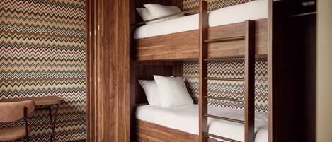 Una cama litera con estructura de madera y ropa de cama blanca, una pequeña mesa con una silla, y una pared con diseño.