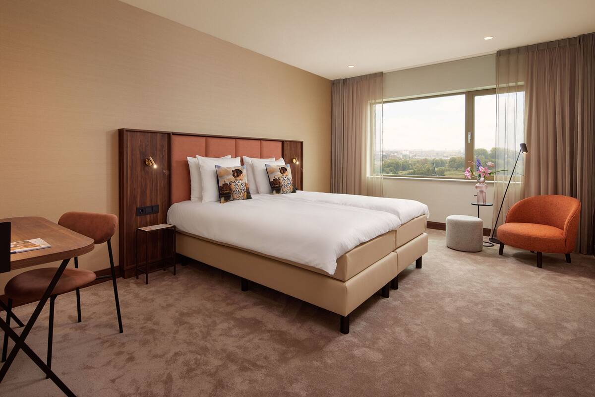 Een hotelkamer met een groot bed, een bureau met stoel, een klein tafeltje en uitzicht op de stad door het raam.