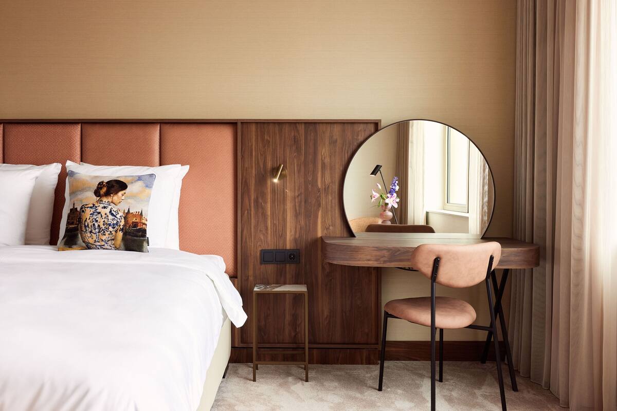Een hotelkamer met een bed, een klein bureau met stoel, een ronde spiegel en een nachtkastje met lamp.