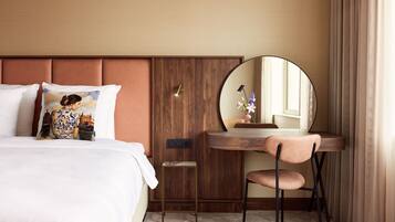 A hotel room with a bed, a small desk with a chair, a round mirror, and a nightstand with a lamp.