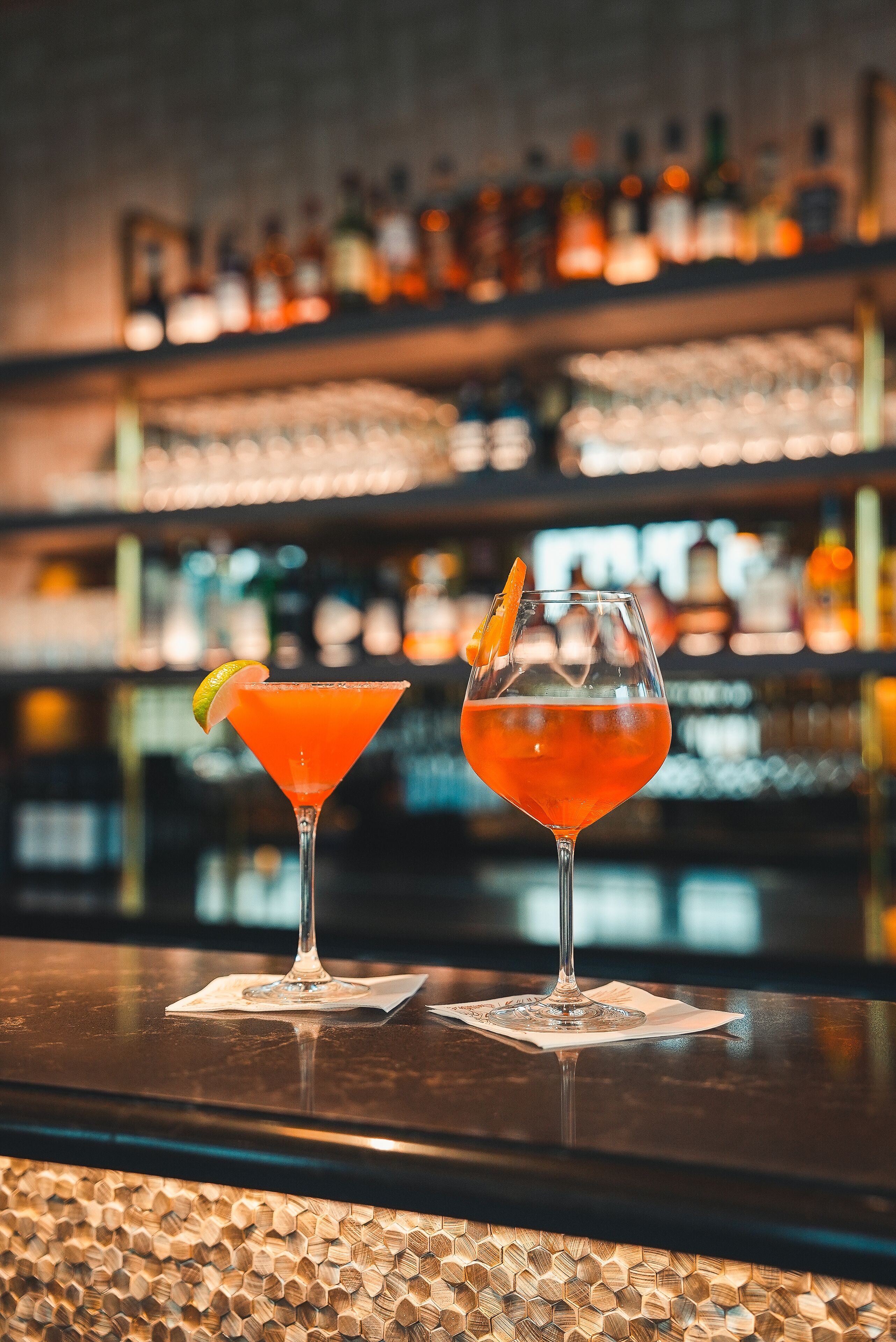 Deux cocktails sur le comptoir d’un bar, avec en arrière-plan, flou, des étagères remplies de bouteilles.