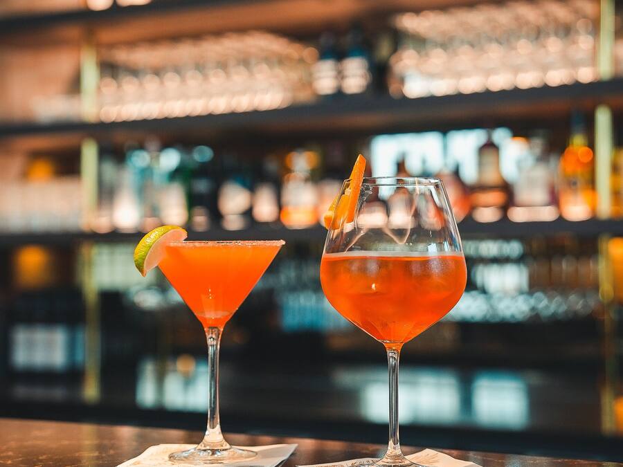 Two cocktails on a bar counter with a blurred background of shelves stocked with bottles.