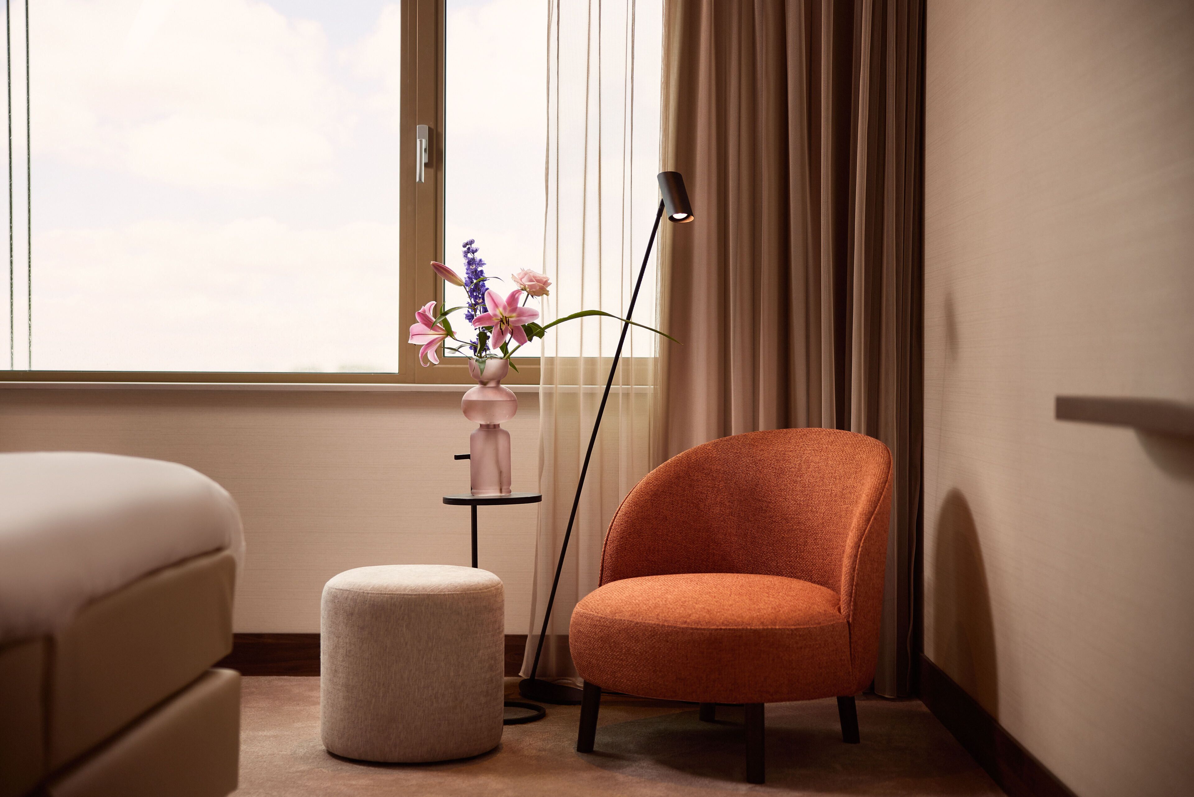 A modern room with a large window, a standing lamp, a pink vase with flowers, a round ottoman, and a plush armchair.