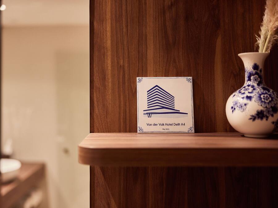 A wooden reception desk with a sign for Van der Valk Hotel Delft A4, a blue and white vase with dried pampas grass, and a blurred background suggesting a hotel interior.