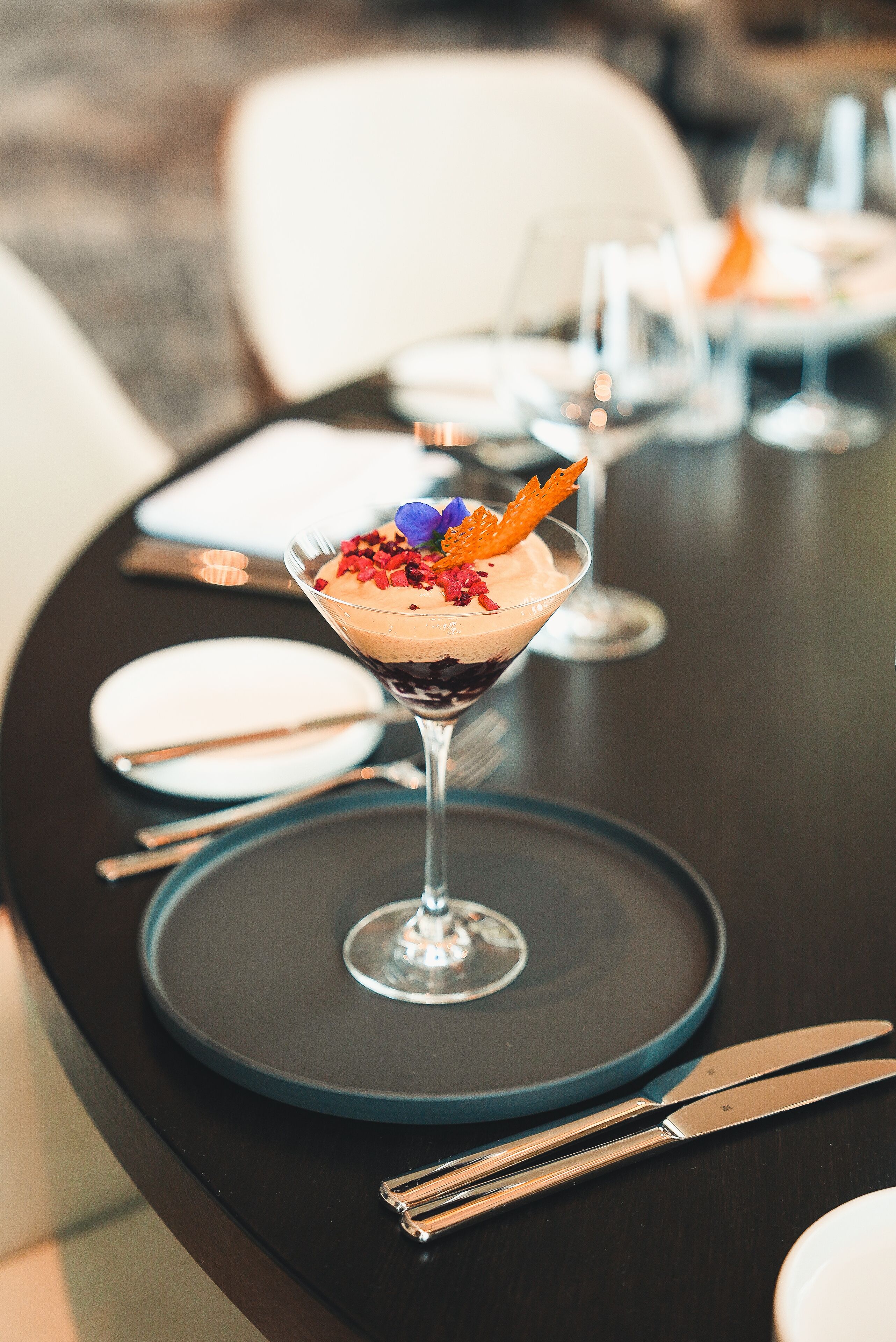 A dessert in a martini glass on a table with a dark tablecloth, silverware, and a white plate.