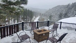A snow-covered deck with a stone fire pit and metal chairs, overlooking a forested valley.