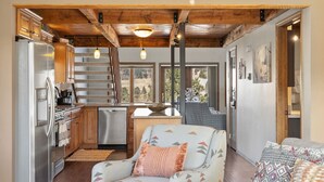 A compact kitchen with a refrigerator, stove, and dishwasher. There is a dining area with a table and chairs, and a sofa with patterned cushions. The room has wooden beams on the ceiling and large windows.