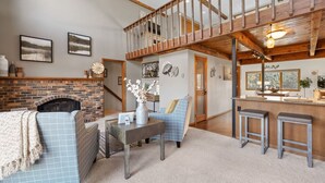 A spacious living area with a fireplace, a high ceiling with exposed beams, a dining area with bar stools, and a staircase leading to an upper level.