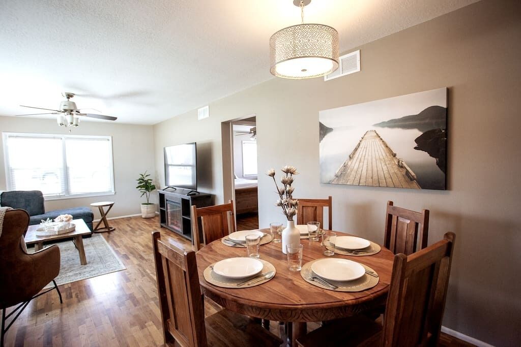 Un coin repas avec une table en bois dressée pour quatre personnes, entourée de chaises en bois, d’un ventilateur de plafond et d’un grand tableau représentant une jetée accroché au mur.