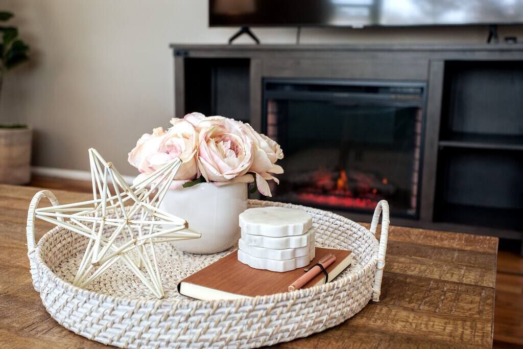 A decorative arrangement on a woven tray with a star-shaped object, a vase with pink flowers, a stack of white candles, and a notebook with a pen.