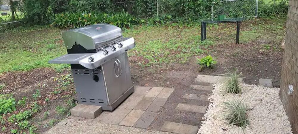 A portable gas grill with a lid, placed on a gravel surface with stepping stones leading to it.