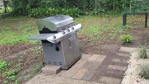 Een draagbare gasgrill met deksel, geplaatst op een grindpad met staptegels ernaartoe.