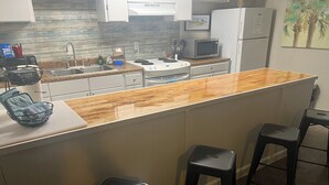 Een keuken met een houten aanrecht, een spoelbak, een magnetron en een koelkast. Er staan twee krukken en een mandje op het aanrecht.