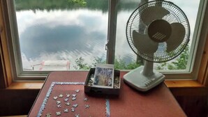 Een raam met uitzicht op een meer en bomen, een puzzel op een tafel, en een staande ventilator.