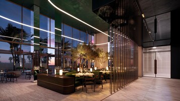 Un espacio interior moderno con mostrador de recepción, zona de estar y ventanas amplias que ofrecen vistas a palmeras y un atardecer.