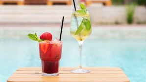 Dos bebidas sobre una mesa de madera al lado de una piscina, con palmeras y sillones de descanso al fondo.