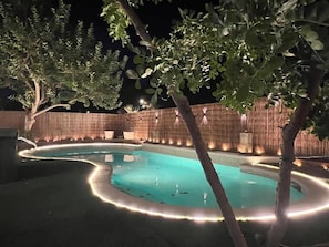 A swimming pool with a curved edge and a lighted border, surrounded by trees and a wooden fence at night. - LUX 6BR/3BA Villa, 10m 2 Strip, Pool+Spa+7 carpark (Henderson)