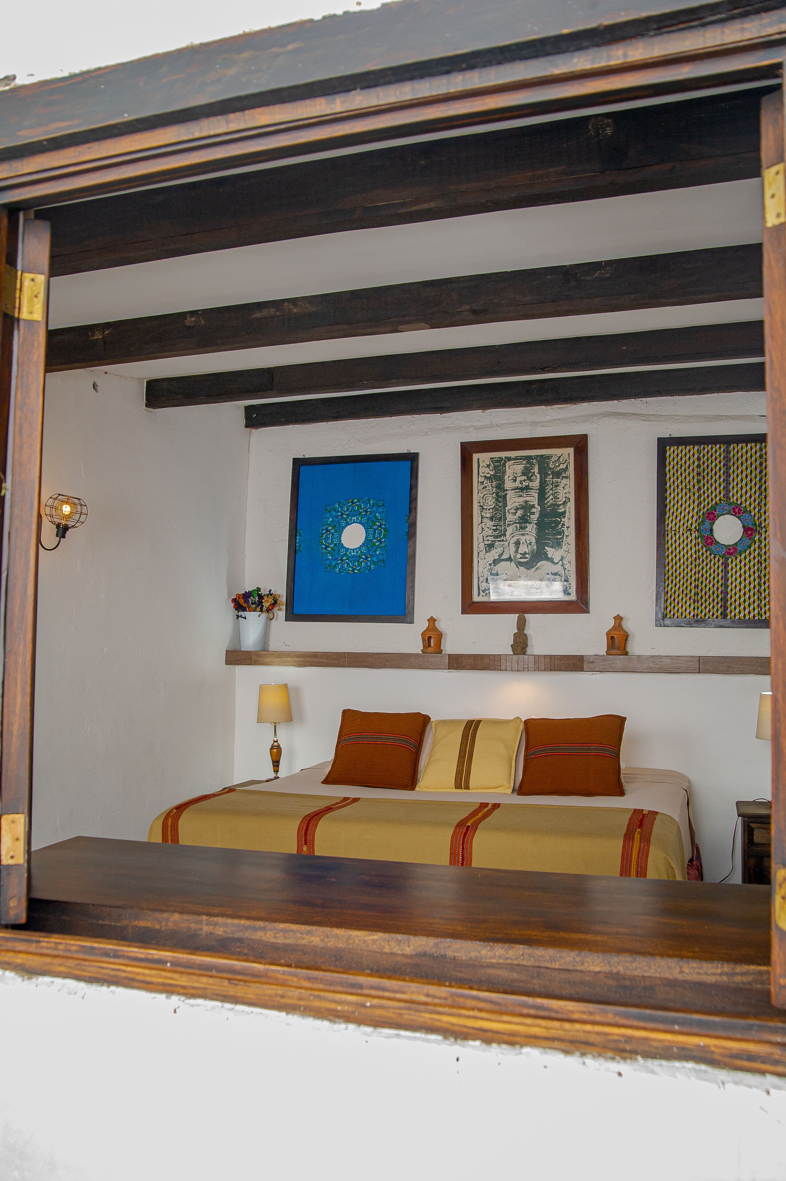 Un dormitorio con cama, mesita de noche, estante en la pared con objetos decorativos y estructura de techo de madera.