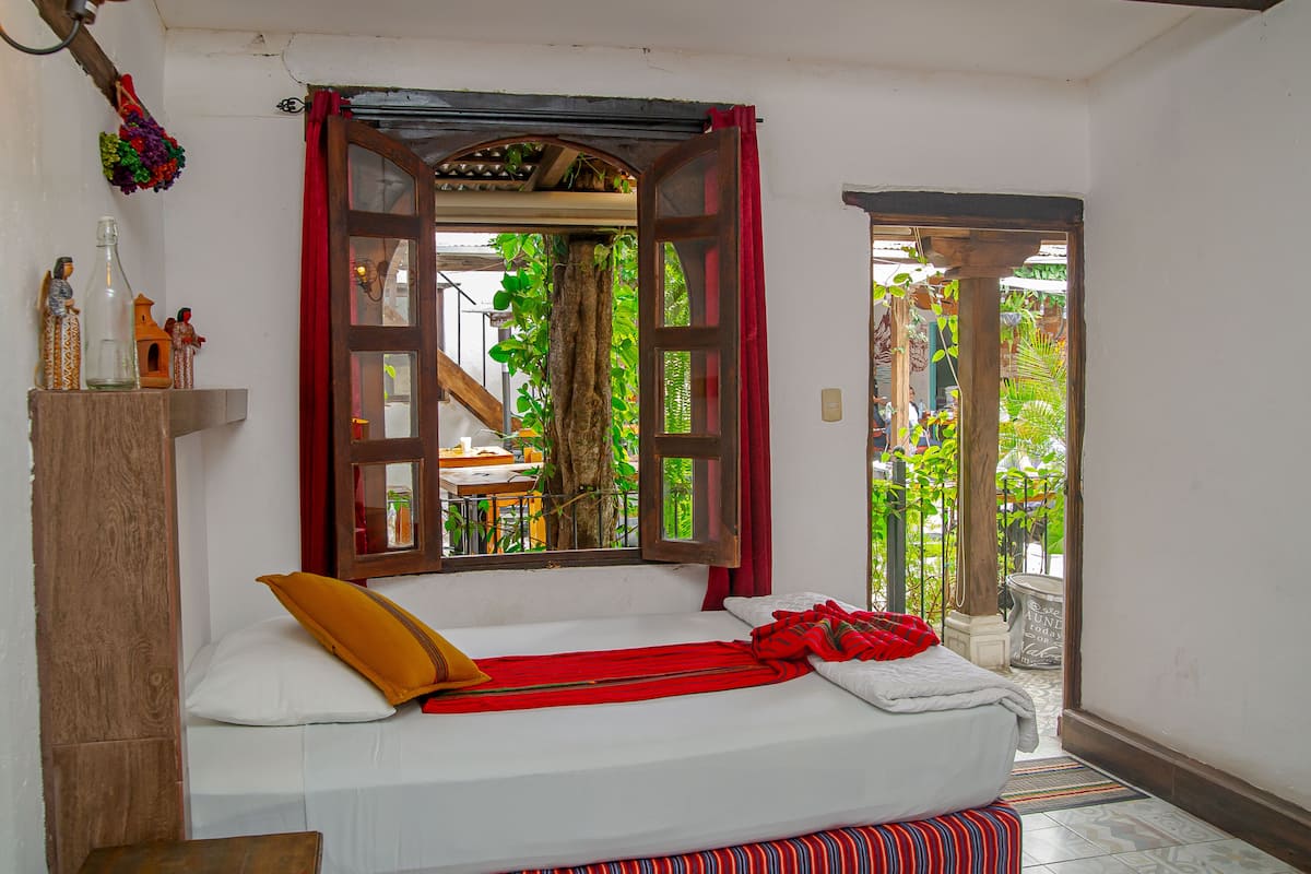 Ein Zimmer mit einem Bett, einem Holzschrank, einem Fenster mit roten Vorhängen und Blick ins Grüne.