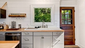 A kitchen with a wooden countertop, white cabinets, a window with a view of greenery, and a wooden door. - The Homestead - Walk to village, bring the pup! (Johnson)
