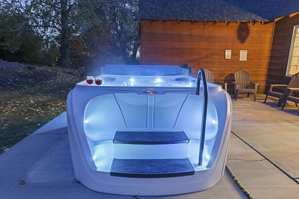 A hot tub with a built-in seat and steps, positioned on a concrete surface near a wooden building with chairs and a window.