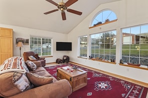 A spacious living room with a large window, a ceiling fan, a television, and a cozy fireplace.