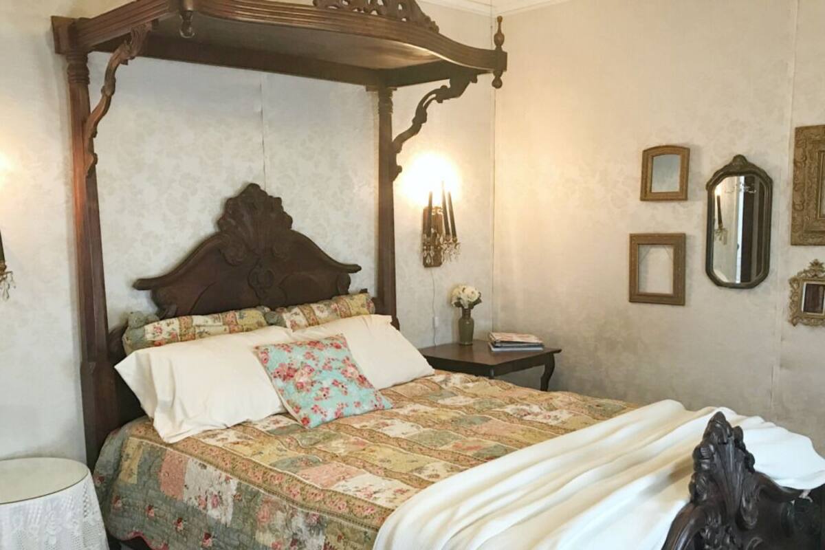 A bedroom with a four-poster bed, a bedside table, a lamp, and wall-mounted mirrors.