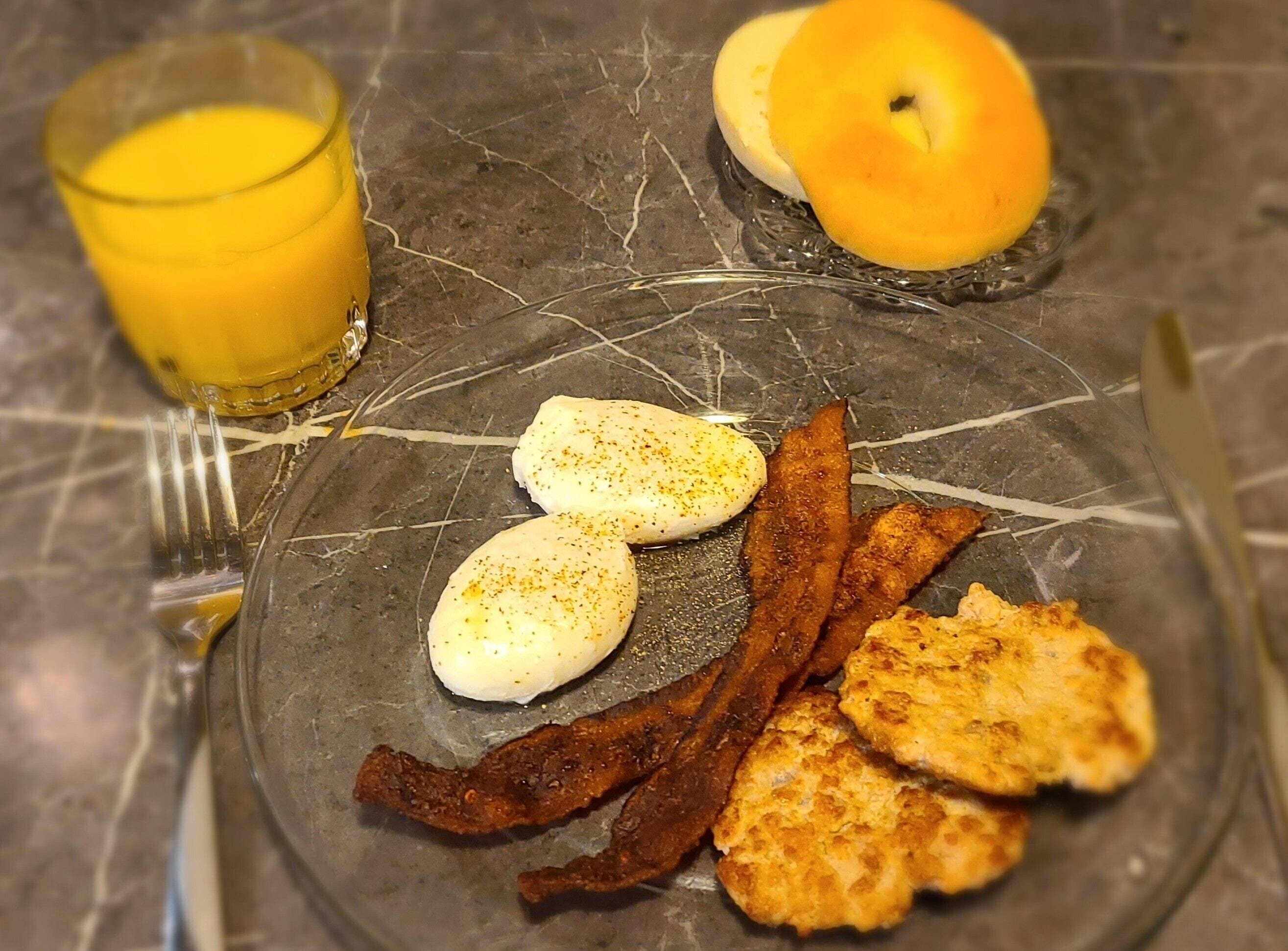 Ein Teller mit Eiern, Speck und Bratkartoffeln, ein Glas Orangensaft und ein Bagel auf einer Marmortischplatte.