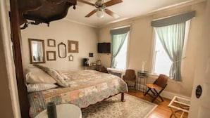 A bedroom with a bed, a chair, a small table, a ceiling fan, and a window with curtains. - The Bissing House (Hays)