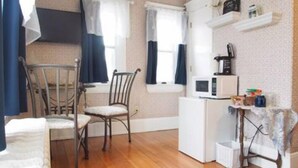 A small, compact room with a wooden floor, a white refrigerator, a microwave, a chair, and a bed. - The Bissing House (Hays)