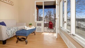 A cozy living room with a sofa, ottoman, and a table with a plant. A sliding glass door leads to a balcony with a view of a snowy landscape. - Great River Road - Victorian Historic Home (1898) (Fountain City)