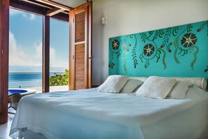 A bedroom with a large bed, white pillows, and a view of the ocean through an open door. - Casa Tiffany - Ilhabela (Ilhabela)