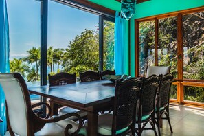A dining area with a large table and chairs, large windows with curtains, and a view of palm trees and greenery. - Casa Tiffany - Ilhabela (Ilhabela)