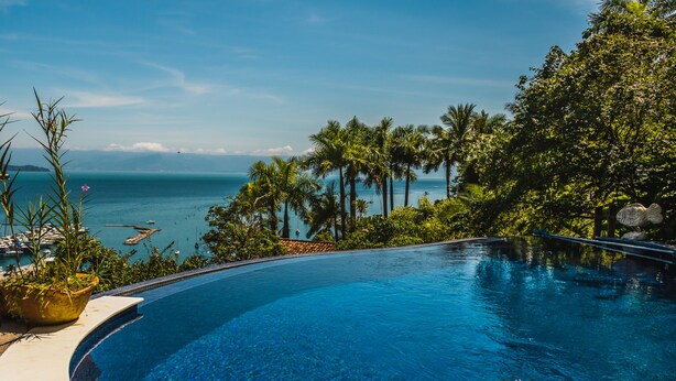 A clear blue infinity pool with a view of the ocean and palm trees. - Casa Tiffany - Ilhabela (Ilhabela)
