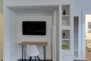 A room with a white brick wall, a flat-screen TV mounted above a wooden desk, a white chair, and a white shelving unit with decorative items.