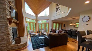 A spacious living room with a stone fireplace, wooden flooring, and large windows offering a view of the outdoors.