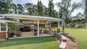 Un patio couvert équipé d’un barbecue et d’un espace salon, entouré d’une pelouse bien entretenue et d’arbres.