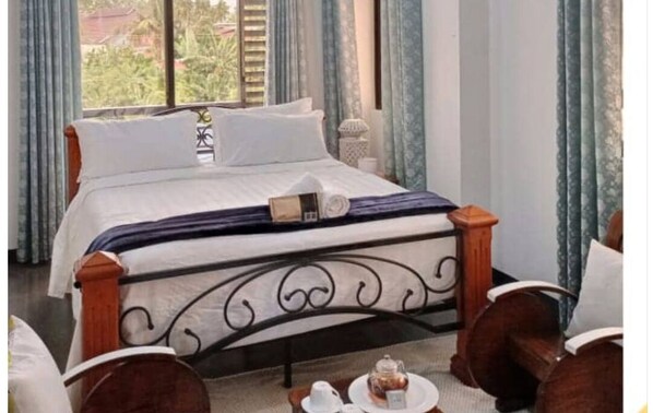 Un lit bien fait, avec une tête de lit sombre, une table de chevet avec une lampe, et une vue sur la verdure à l’extérieur.