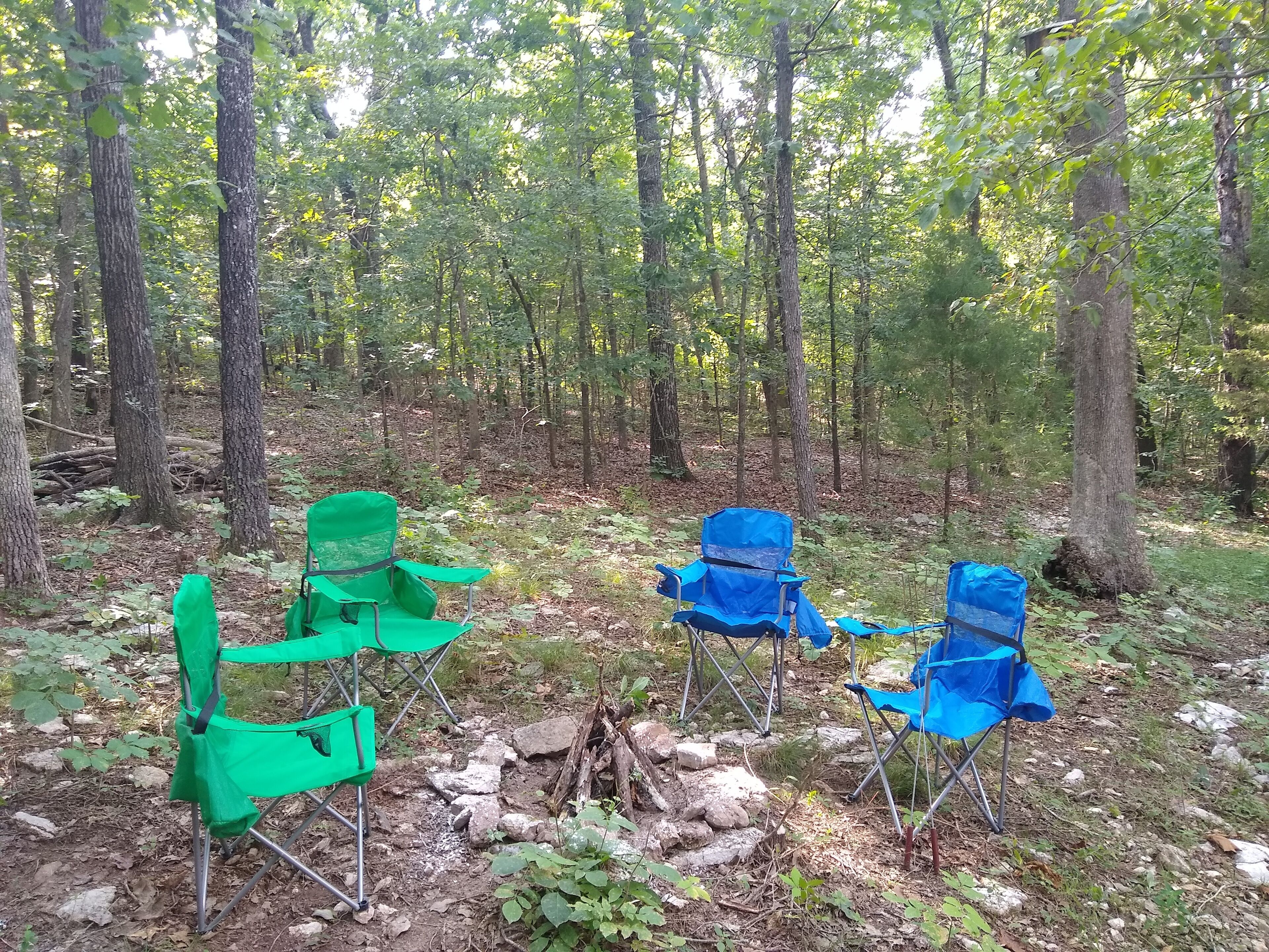 A forest clearing with two green and two blue camping chairs arranged around a small fire pit.