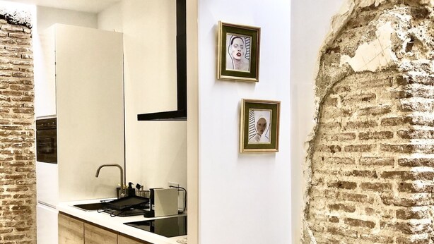 Een moderne keuken met een centraal eiland, een ingebouwde oven en een grote spiegel. Twee ingelijste foto's hangen aan de muur en er staat een kruk naast een bakstenen muur.